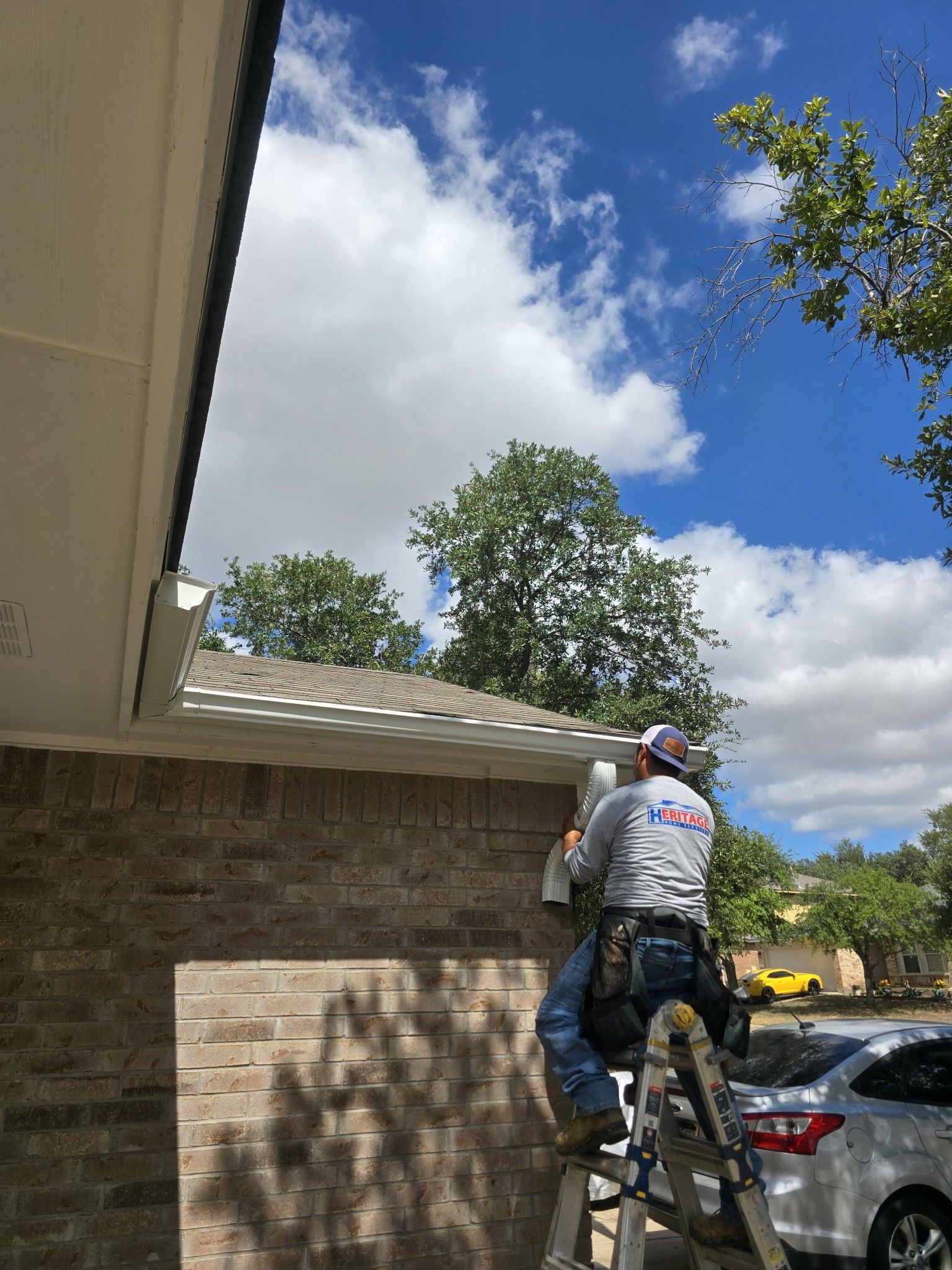 A man on a ladder installs guttering on a brick building under a cloudy sky.