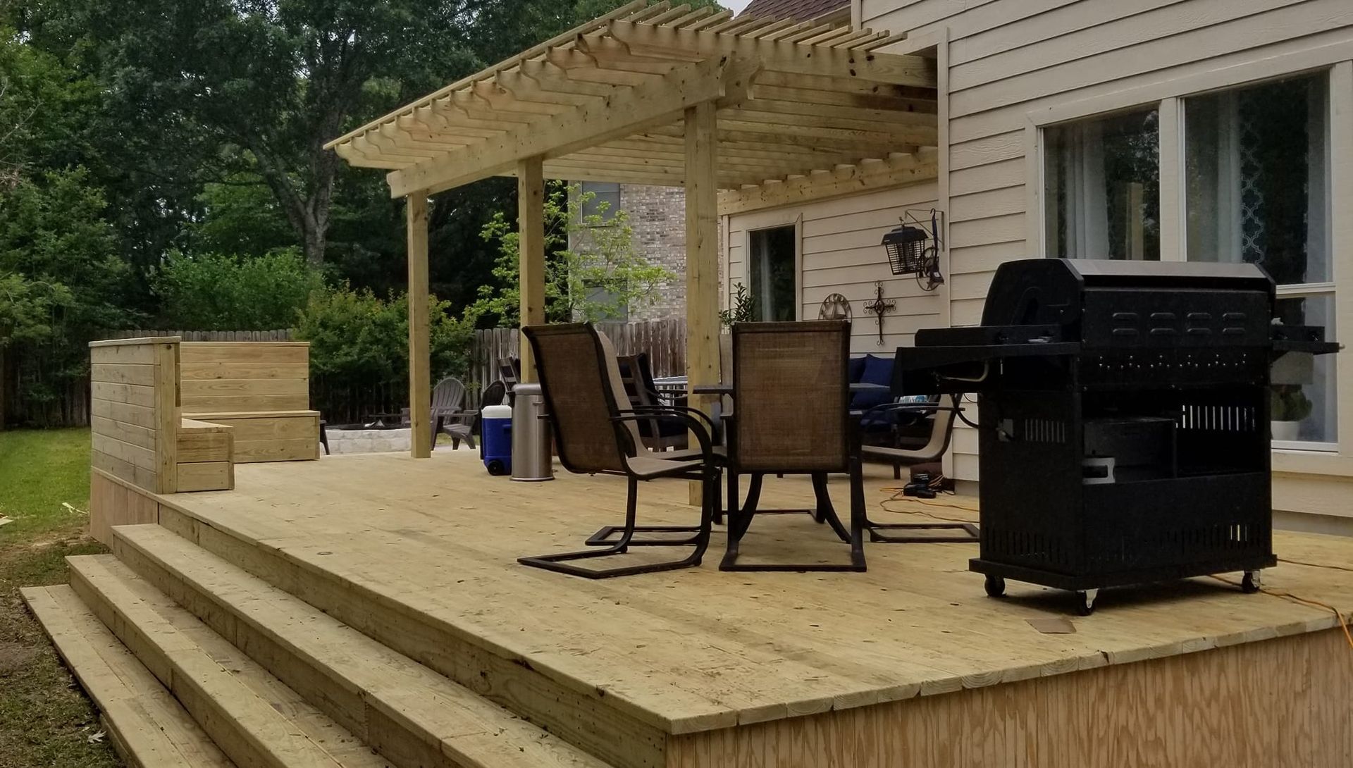 Wooden deck with pergola, grill, table, chairs, and steps leading to a yard near a house.