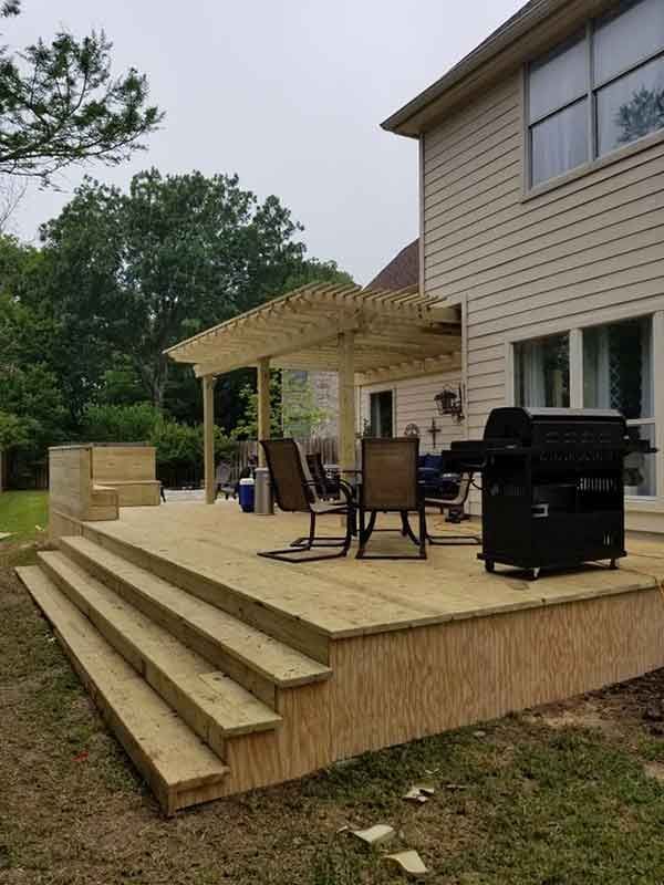 Wooden deck with pergola, stairs, and grill next to a house with beige siding. Overcast sky.