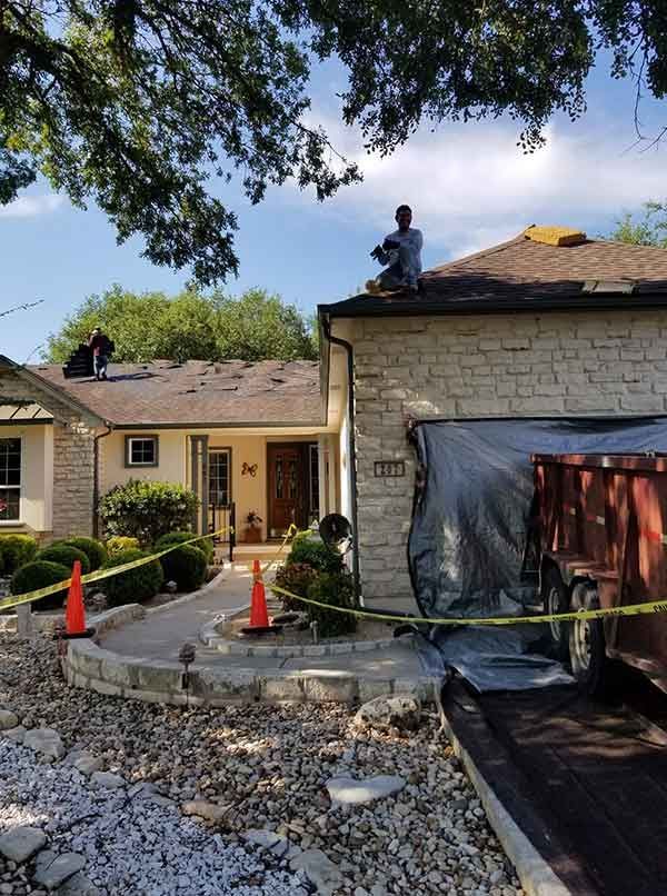 Roofers working on a house; debris, safety cones, and a dumpster are present. Blue sky, sunny day.