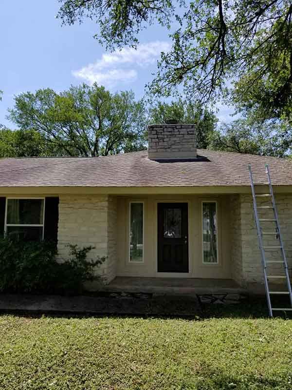Tan house with chimney, ladder, and trees against a blue sky.