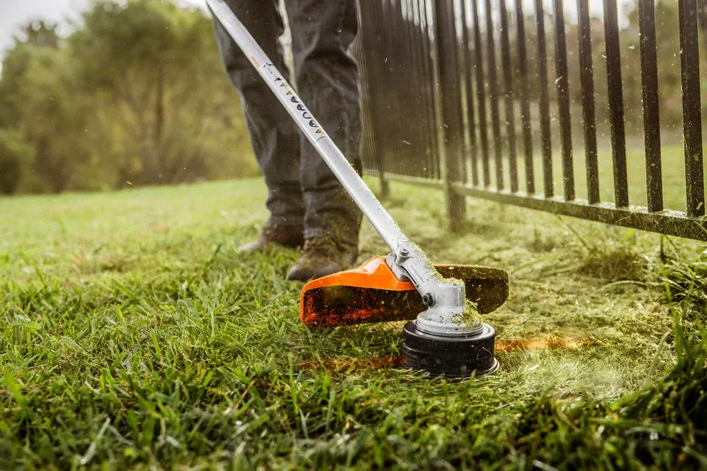 A person is using a lawn mower to cut the grass next to a fence.
