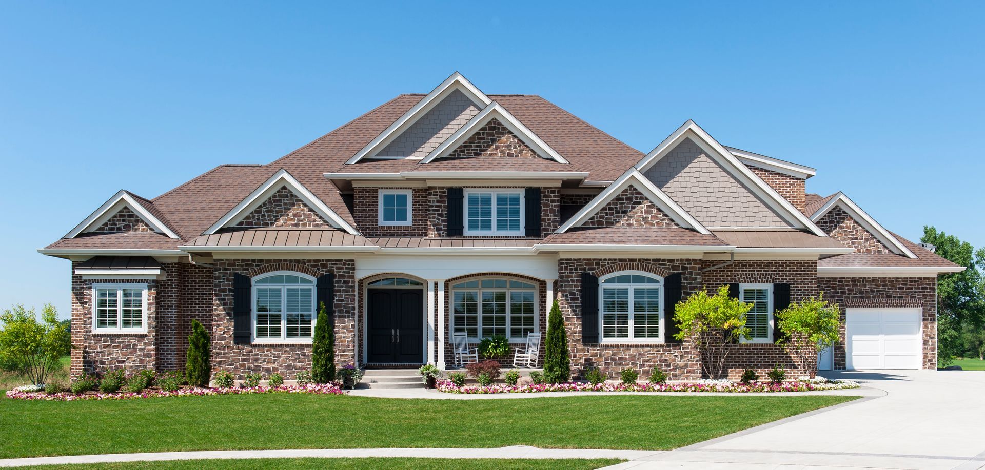 Large brick suburban house with a gray roof, arched windows, and a manicured lawn under a blue sky