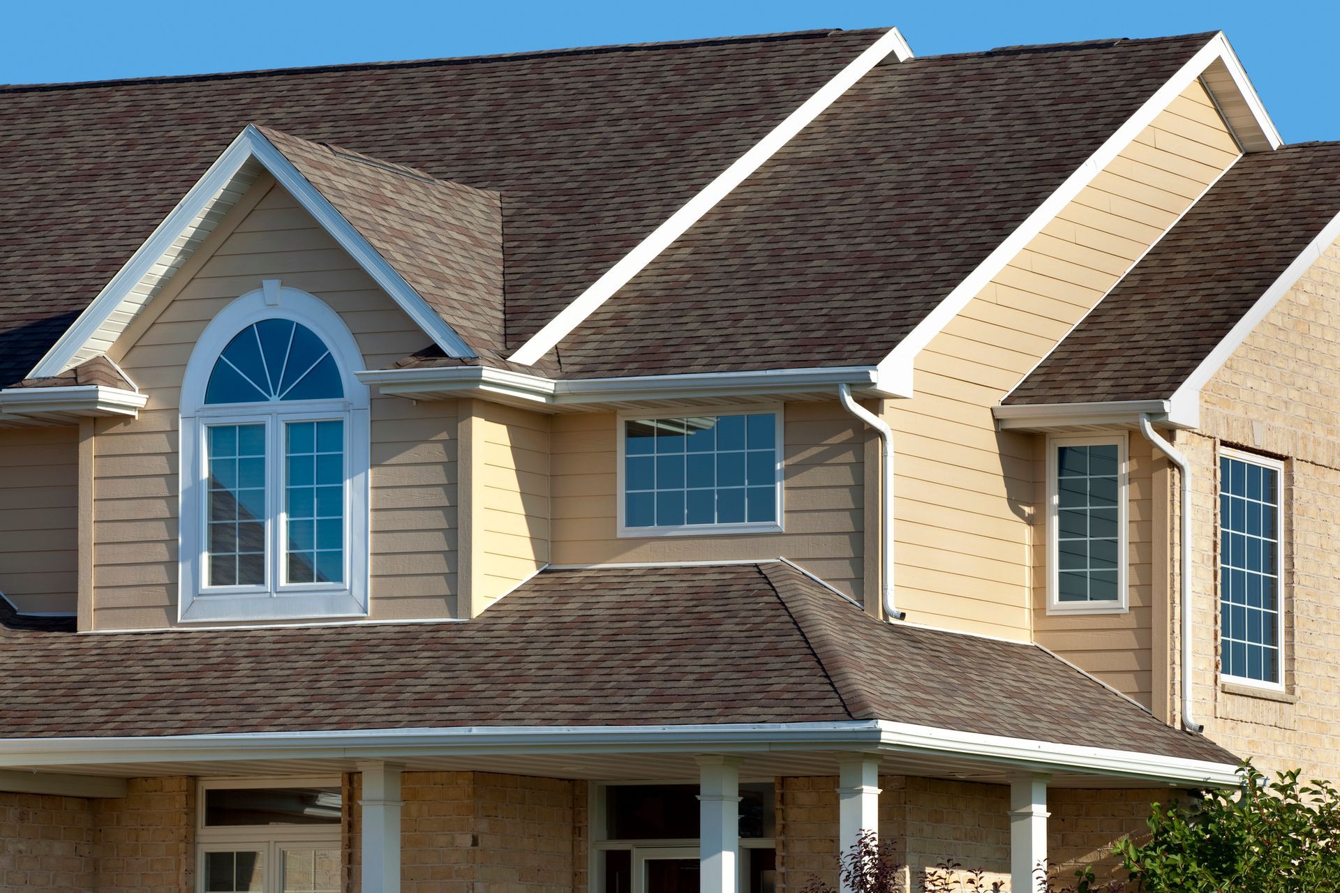 Two-story beige house with brown shingled roof, arched window, and white trim under a blue sky