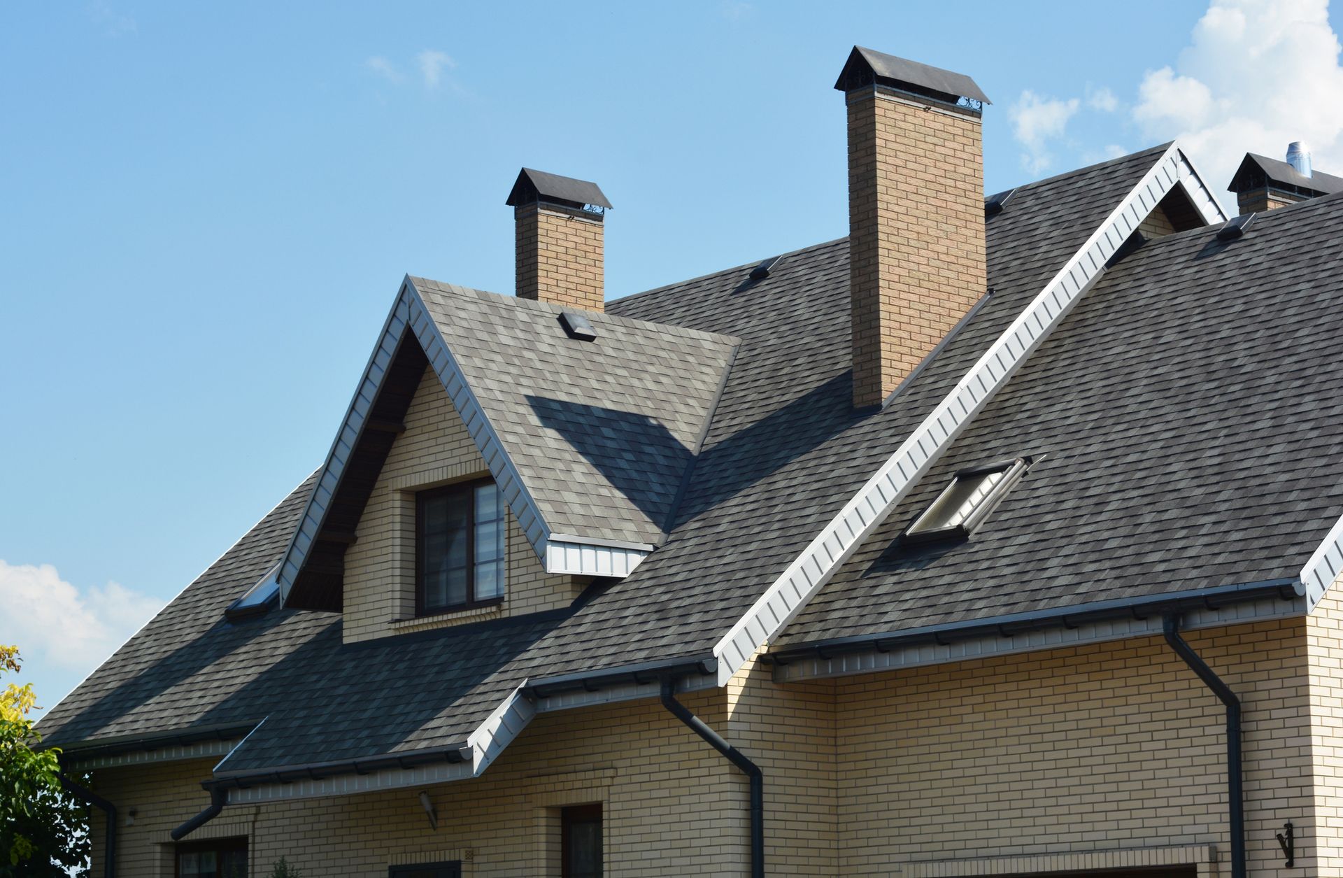 Brick house with steep gray shingle roof and two chimneys under a blue sky