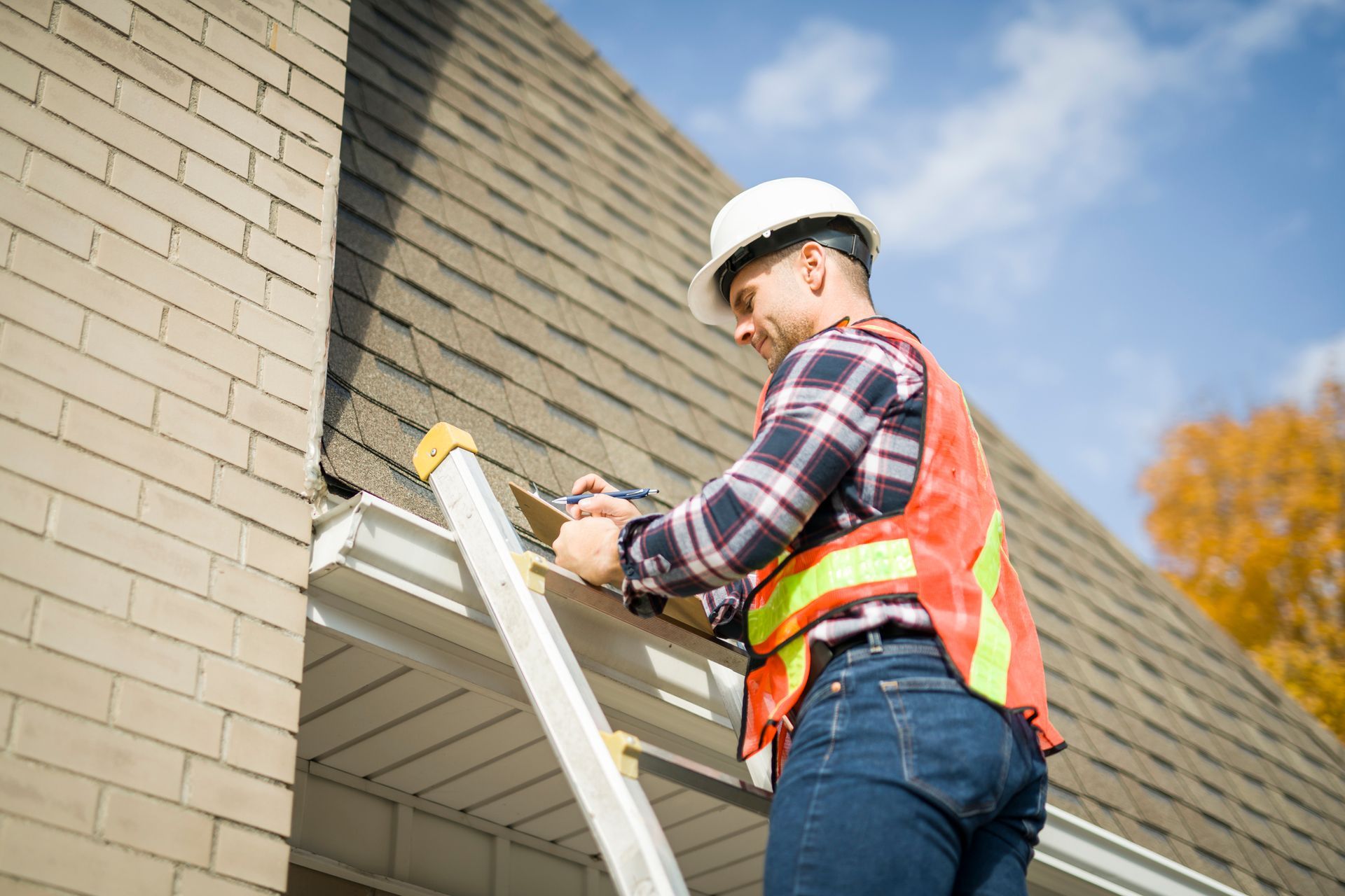 Construction worker on a ladder inspecting a roof with a tool, wearing a hard hat and safety vest.