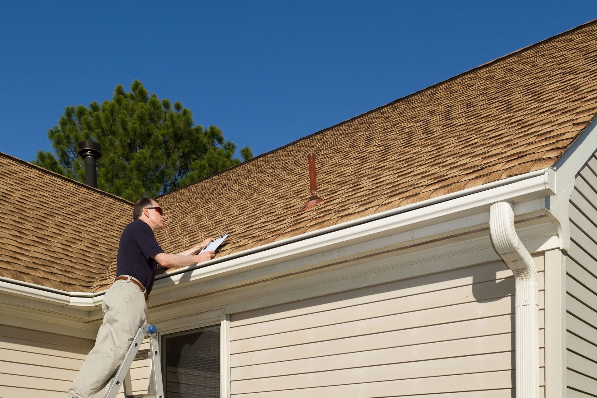 Person on a ladder cleaning gutters on a beige house with a brown shingled roof