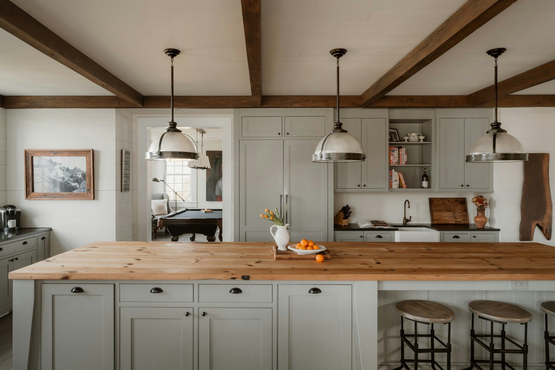 A modern kitchen with a large wooden island, grey cabinets, white walls, and dark pendant lights under exposed wood beams.