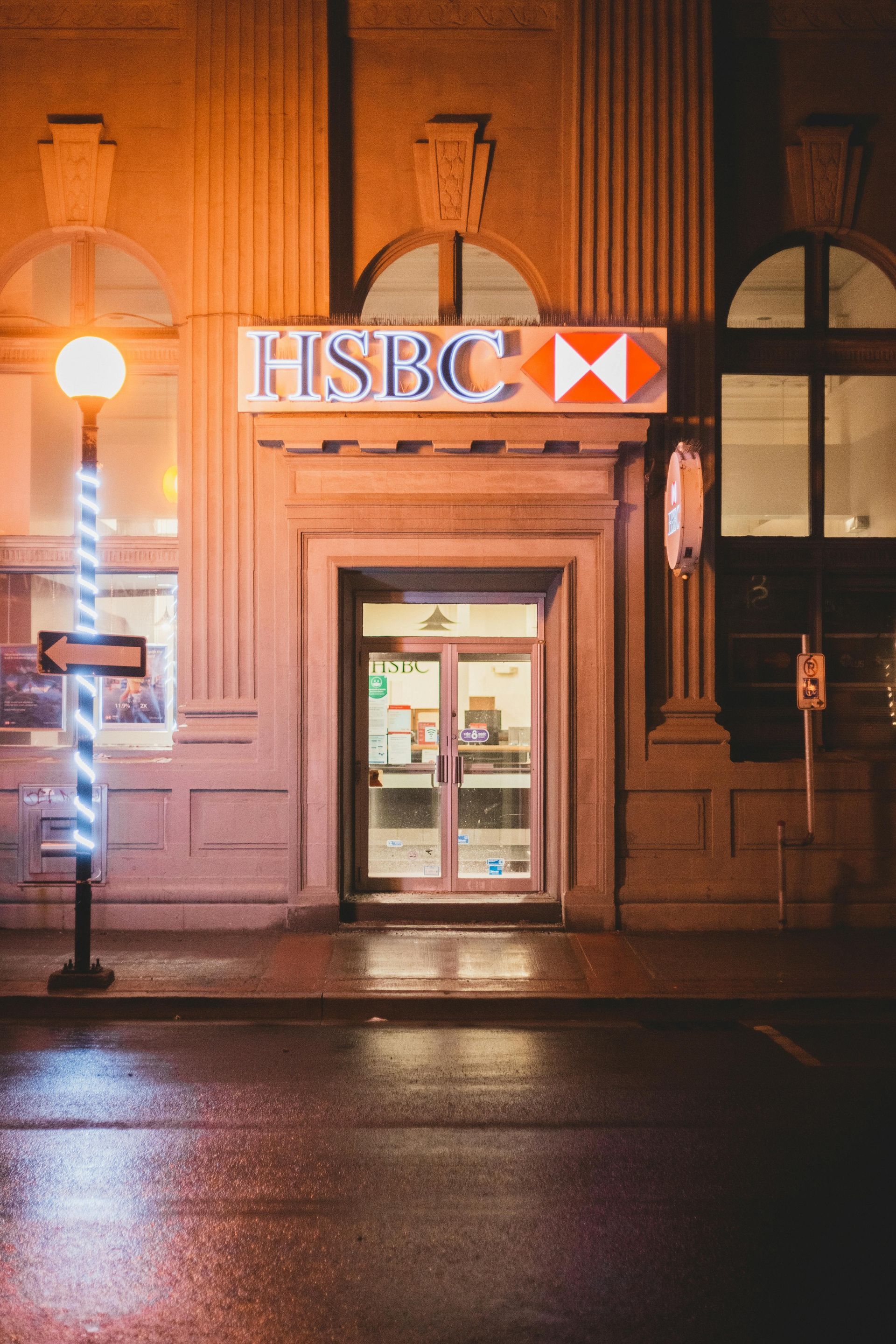 A hsbc bank is lit up at night with a street light in front of it.