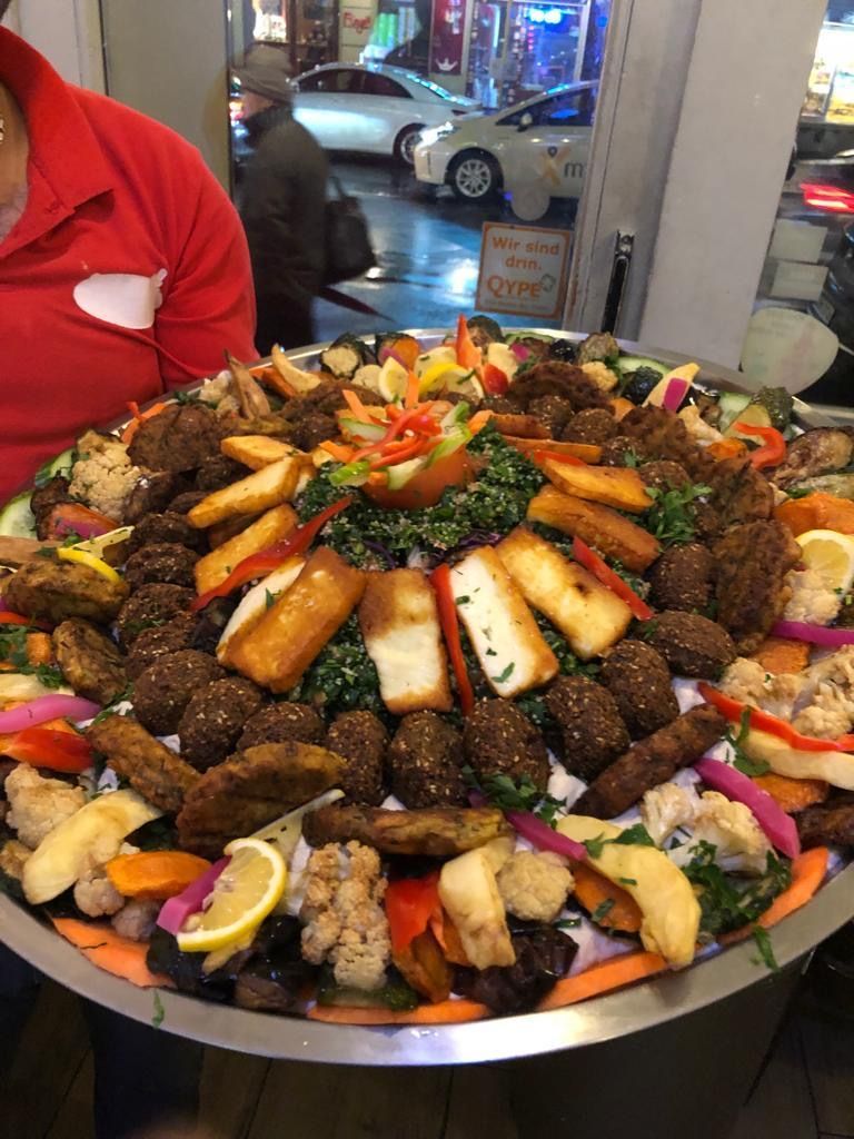 Large platter of falafel, vegetables, and fried halloumi; held by a person in a red shirt, street scene background.