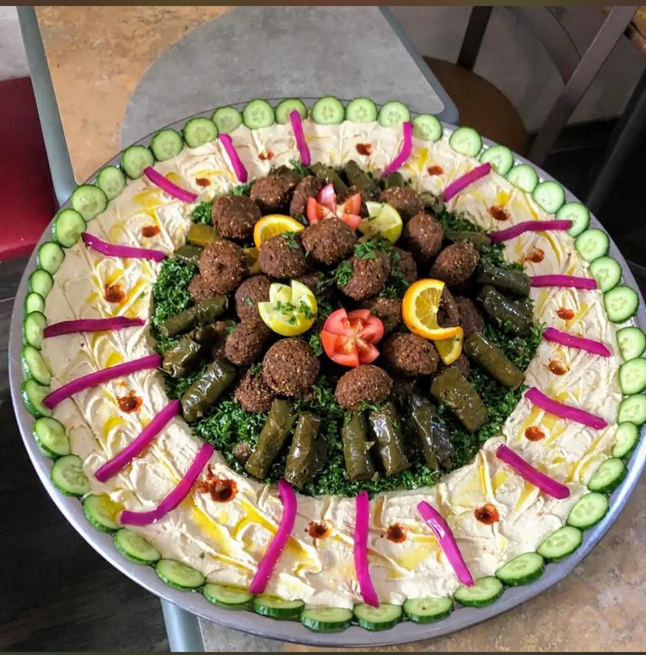 Large platter of hummus with falafel, stuffed grape leaves, pickles, cucumbers, and orange slices.