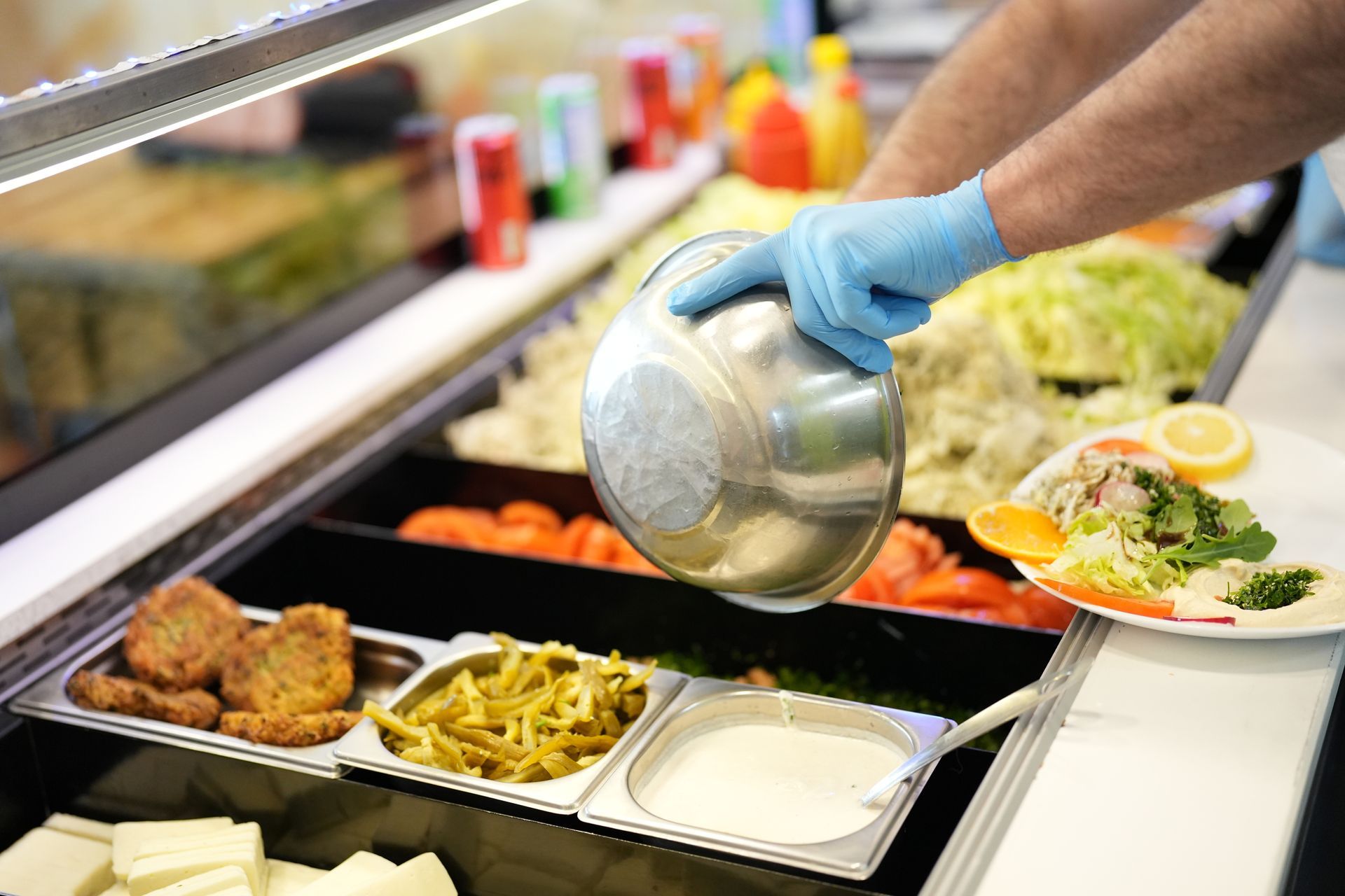 Person preparing food behind a restaurant counter, wearing gloves and using a metal bowl to add ingredients to a salad.