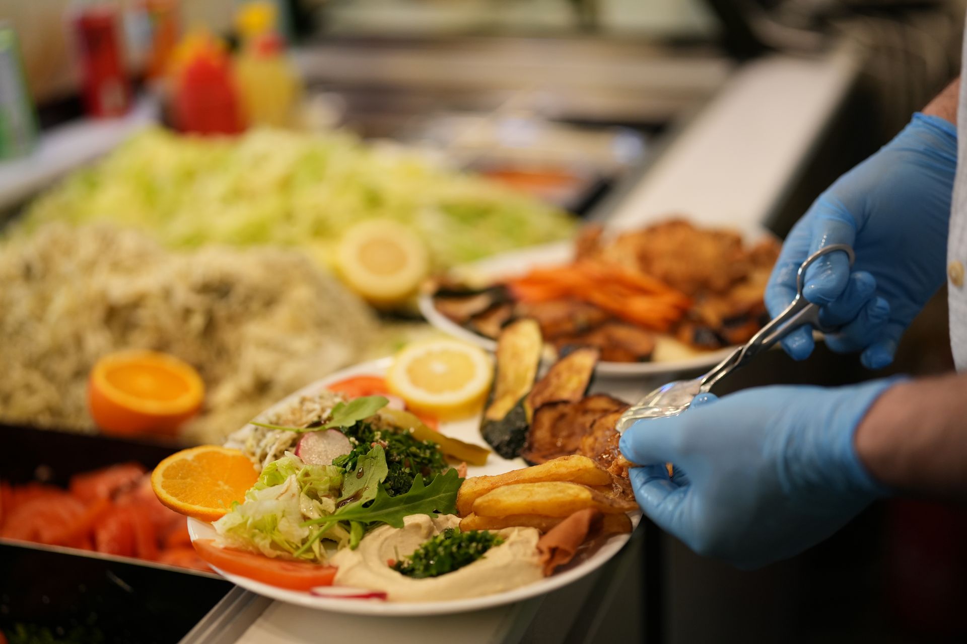 Person in blue gloves preparing a plate of food with salad, hummus, and fried items.