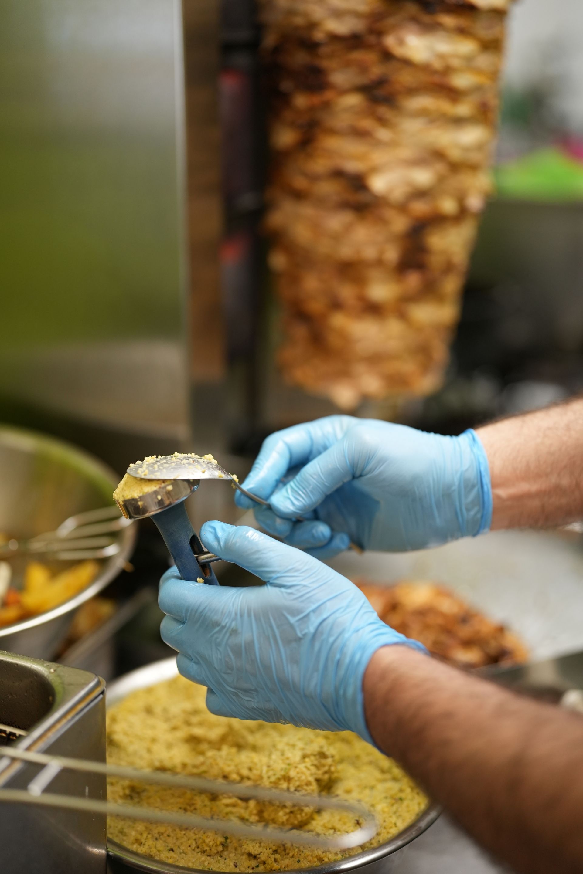 Person in blue gloves preparing food near a rotating shawarma and a plate of yellow grains.