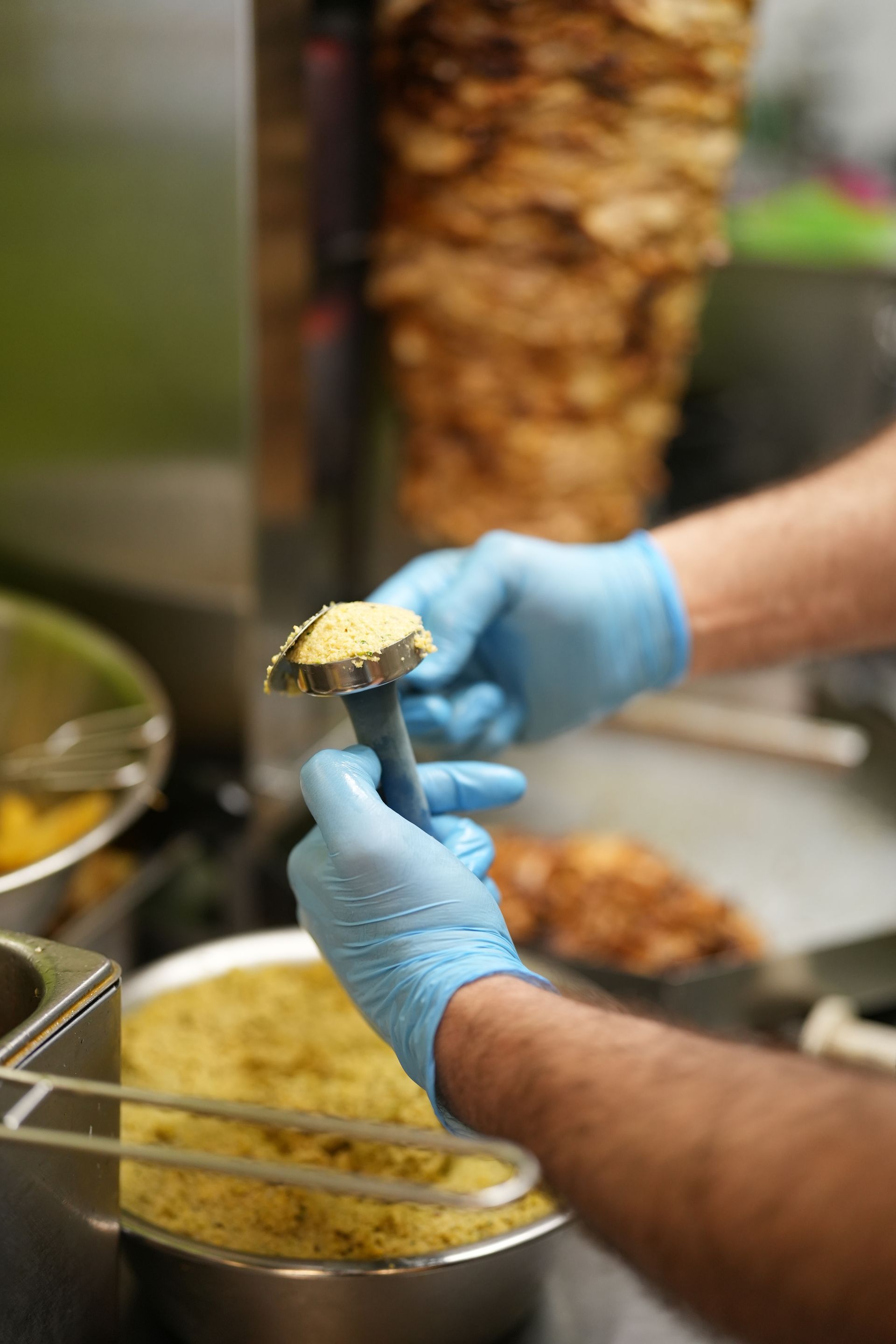 Person in blue gloves using a falafel scoop, shawarma spit in background.