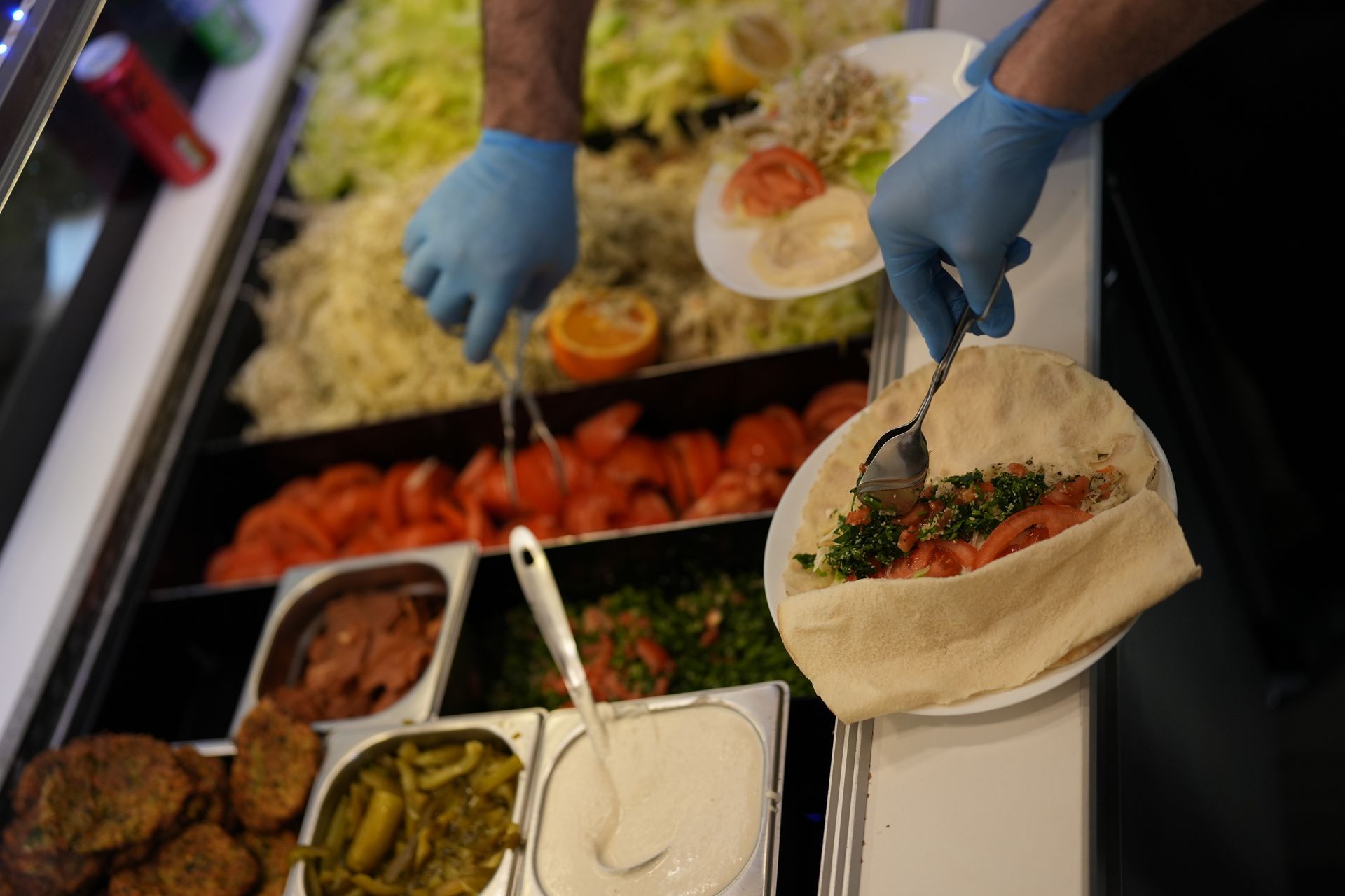 Person preparing a pita sandwich with various toppings, behind a food display case.