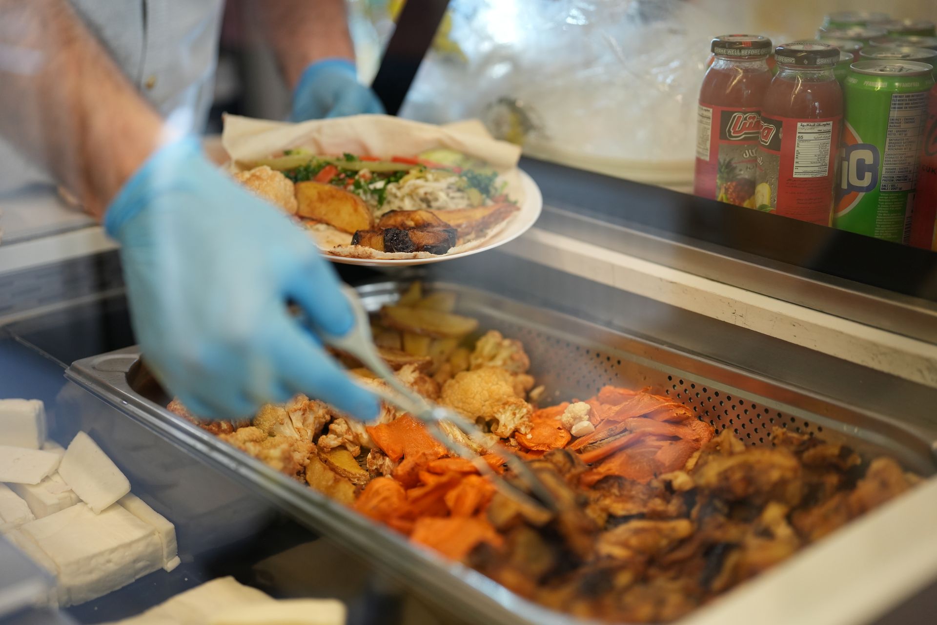 Person wearing blue gloves serving food from a hot bar. Food is visible and a plate is being prepared.