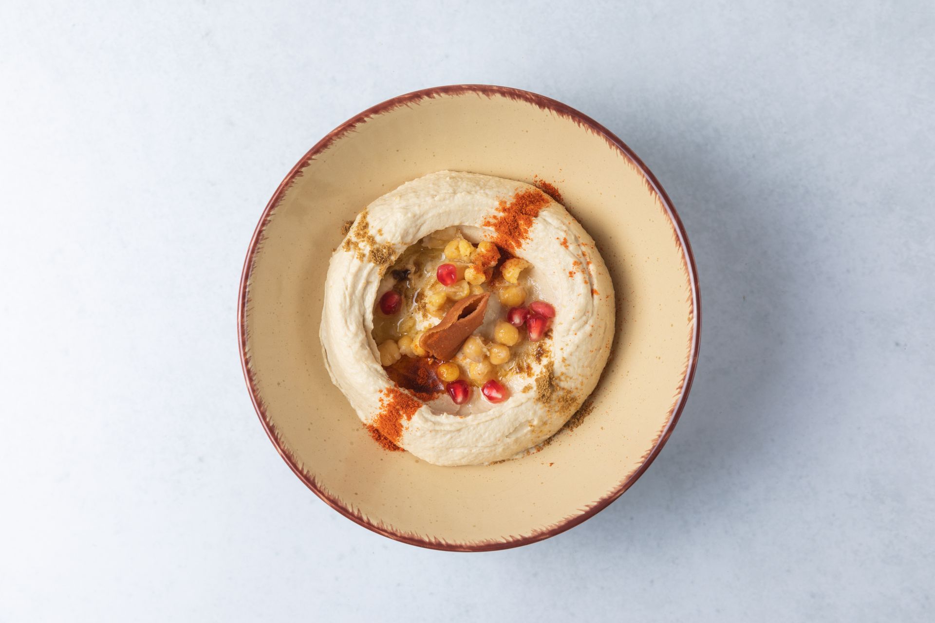 Hummus in a tan bowl, topped with chickpeas, pomegranate seeds, and paprika.