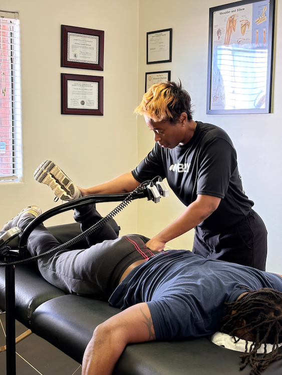 Dr. Natasha Williams assisting a patient lying on a treatment table with a device. Indoors, certificates on the wall.