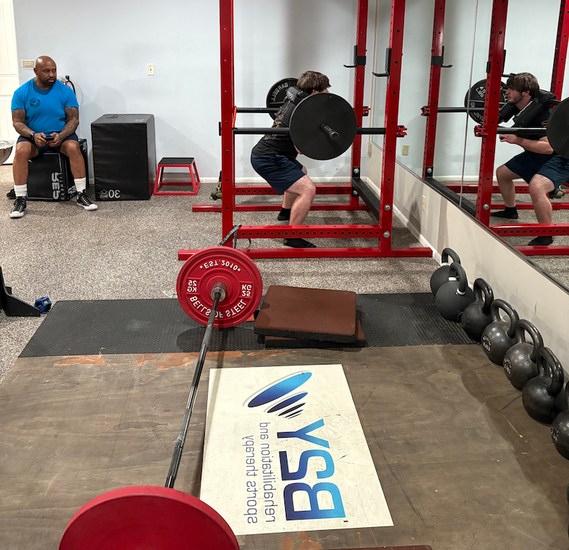 Person squatting with barbell in a gym, red power rack, weight plates, and spotter.