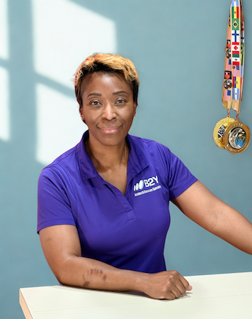 Dr. Natasha Williams of B2Y in purple shirt, smiling, by table; medals in background.