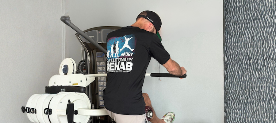 Man in a black shirt exercises on a machine in a room with white walls, near a textured wall.