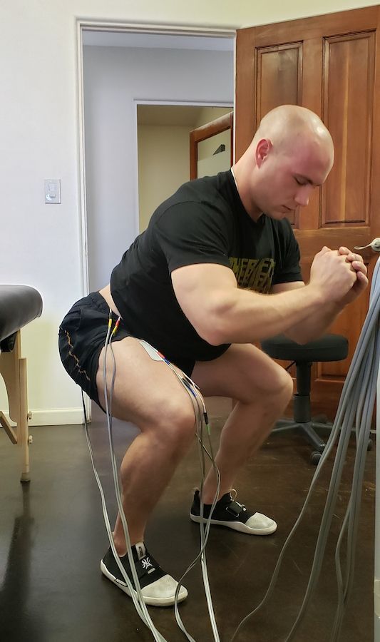 Man in athletic stance, electrodes attached, eyes down, in a squat. Dark shirt, shorts, sneakers. Indoor setting.