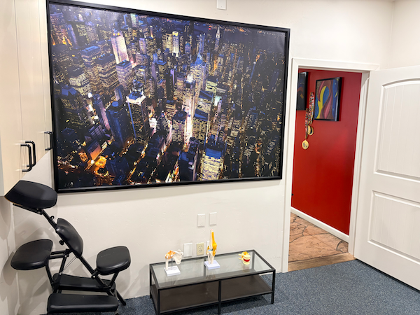Massage chair in a room with a large cityscape photo, small glass table, and a door to a red-walled space.