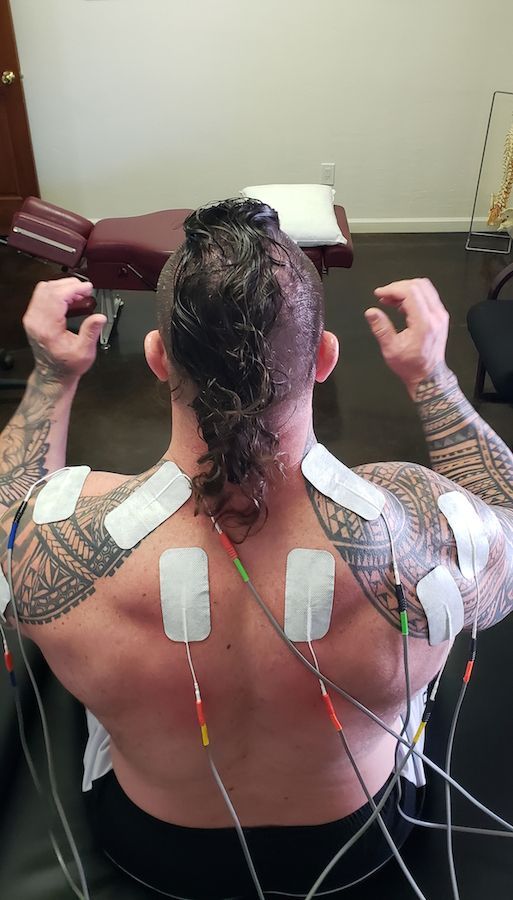 Man with tattoos, mohawk, and electrical stimulation pads on his back, seated in a clinical setting.