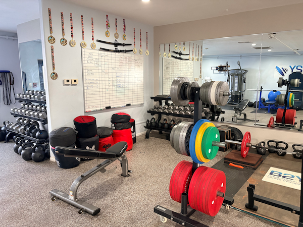 Weight room with weights, bench, mirror, whiteboard, and medals on the wall.