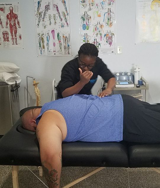 Person receiving back massage on a massage table. Dr. Natasha Williams uses elbow. Clinic setting with anatomical charts.