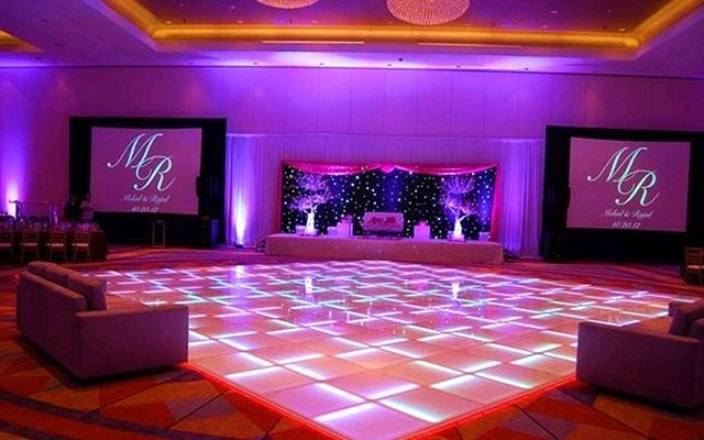 Lit dance floor in a ballroom, with stage, screens showing initials, and seating. Purple and orange lighting.