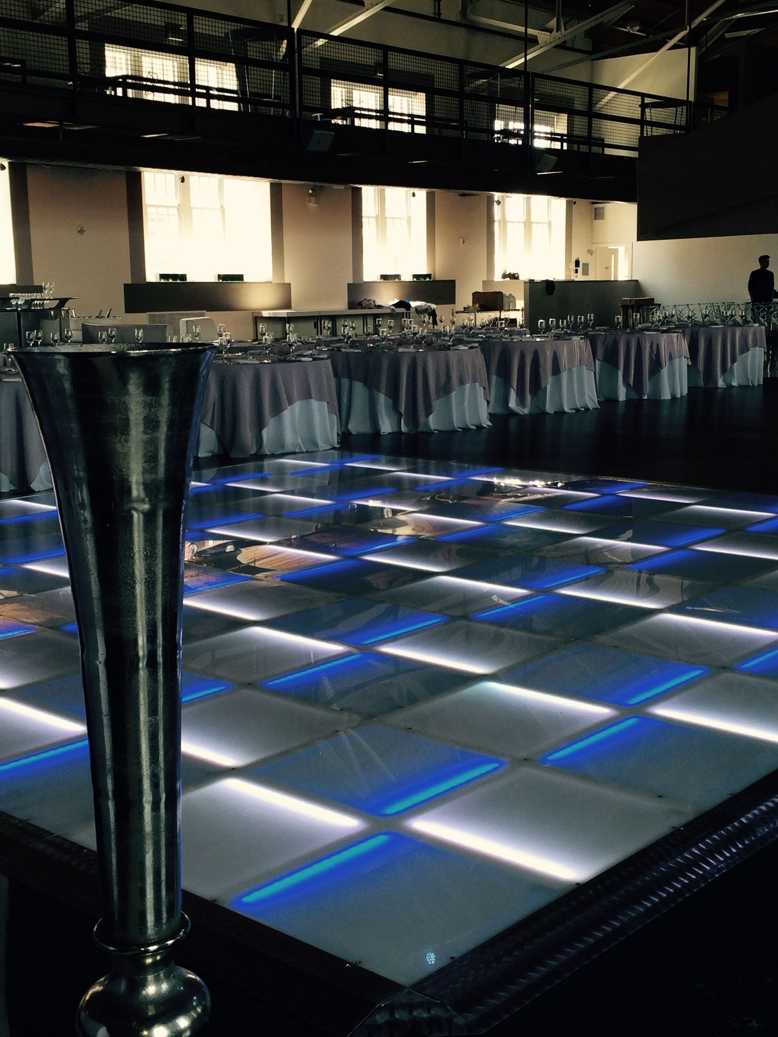 Lighted dance floor in a large venue with tables, a balcony, and a decorative pole in the foreground.