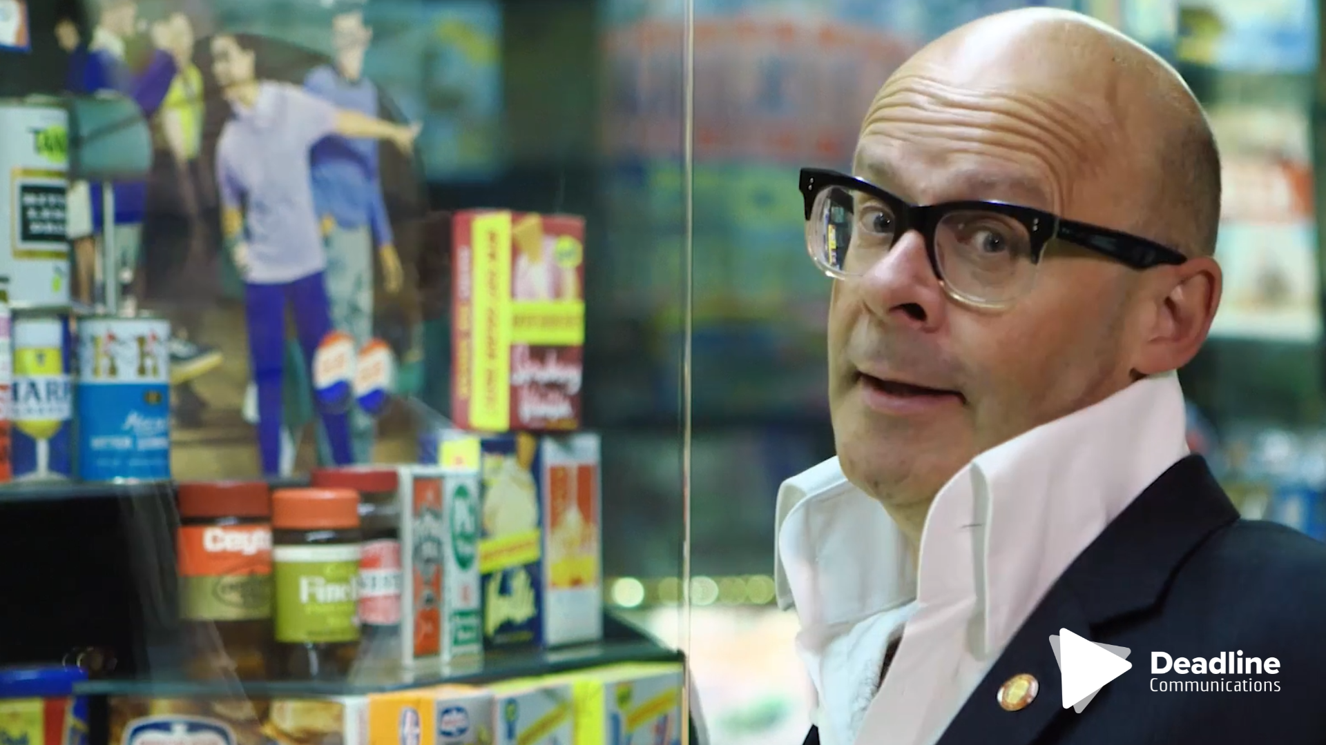 Man with glasses, looking at a display case with various products. He wears a suit jacket and white collared shirt.