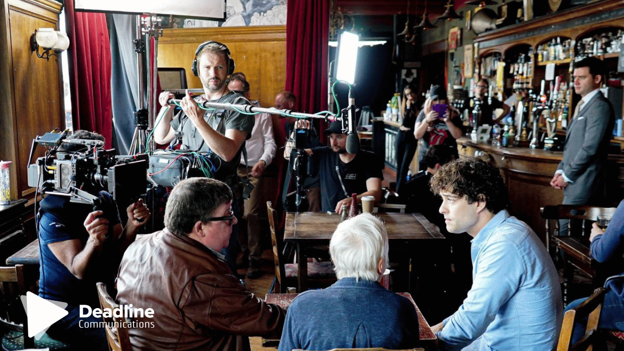 Film crew shooting a scene in a pub. Actors sit at a table; camera crew surrounds them.