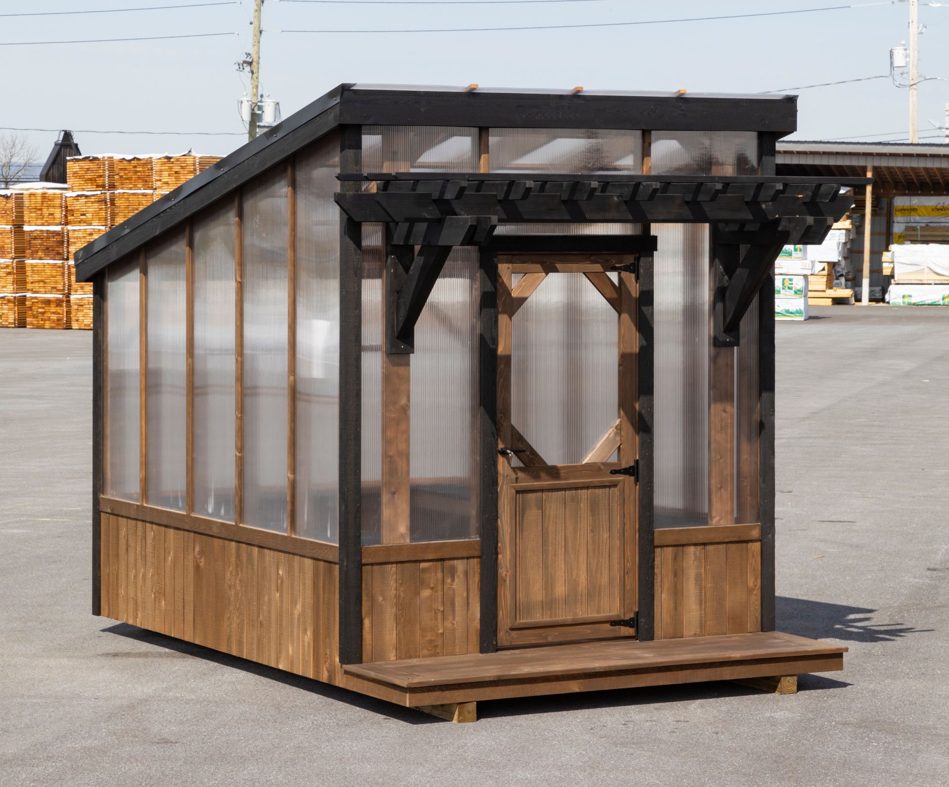 A small, modern wooden greenhouse with a sloped roof, clear panels, a front door, and a decorative pergola, in a lot.