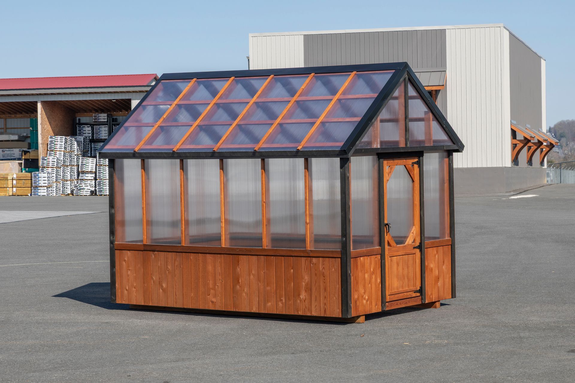 A wooden greenhouse with a gabled roof and translucent panels, standing outdoors on a paved lot.