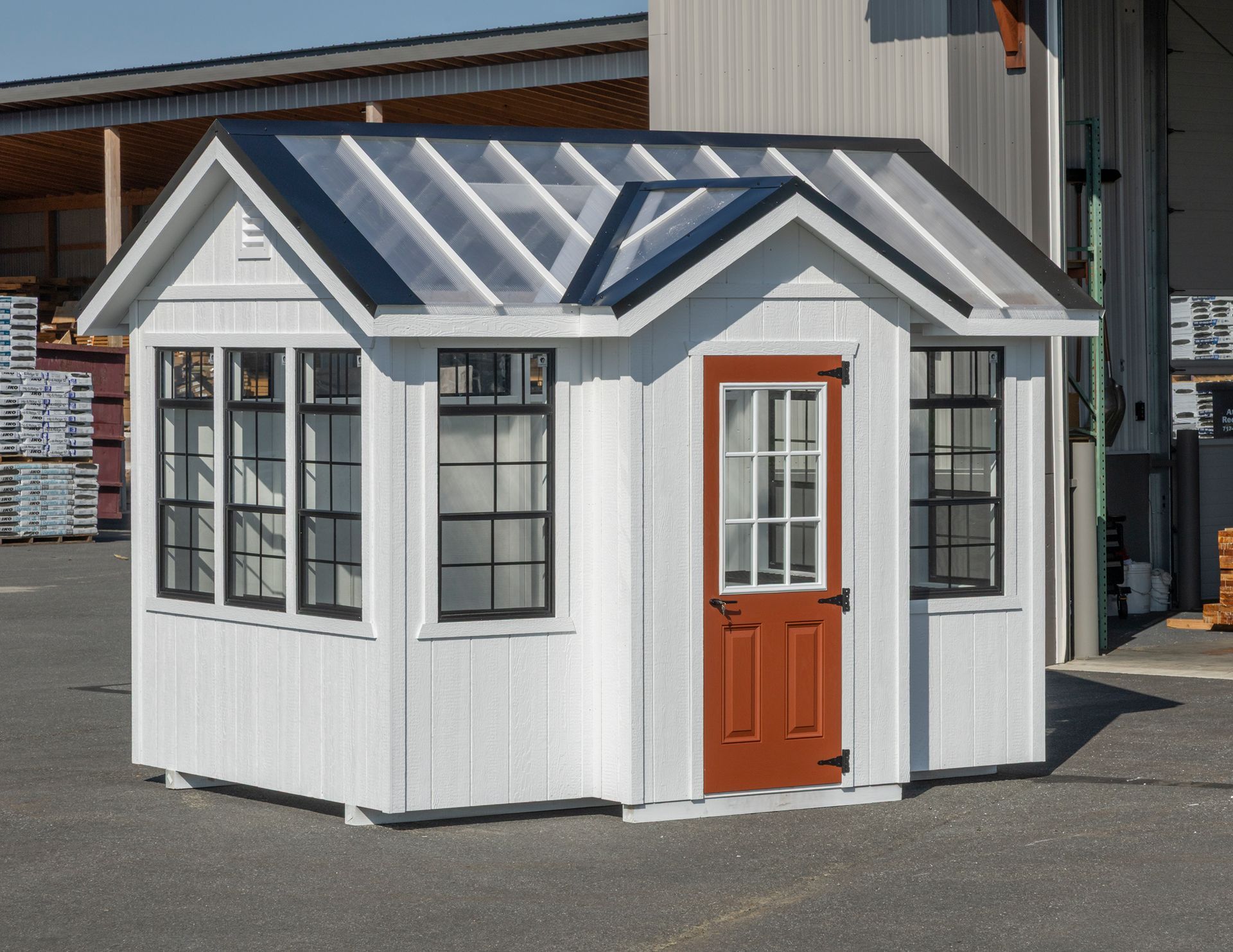 A white shed with a glass roof, black-trimmed windows, and an orange door sitting on a gravel lot.