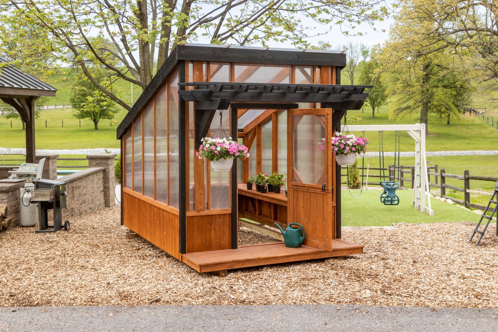 A small, wooden greenhouse with translucent panels, a black frame, and a front porch, set in a backyard landscape.