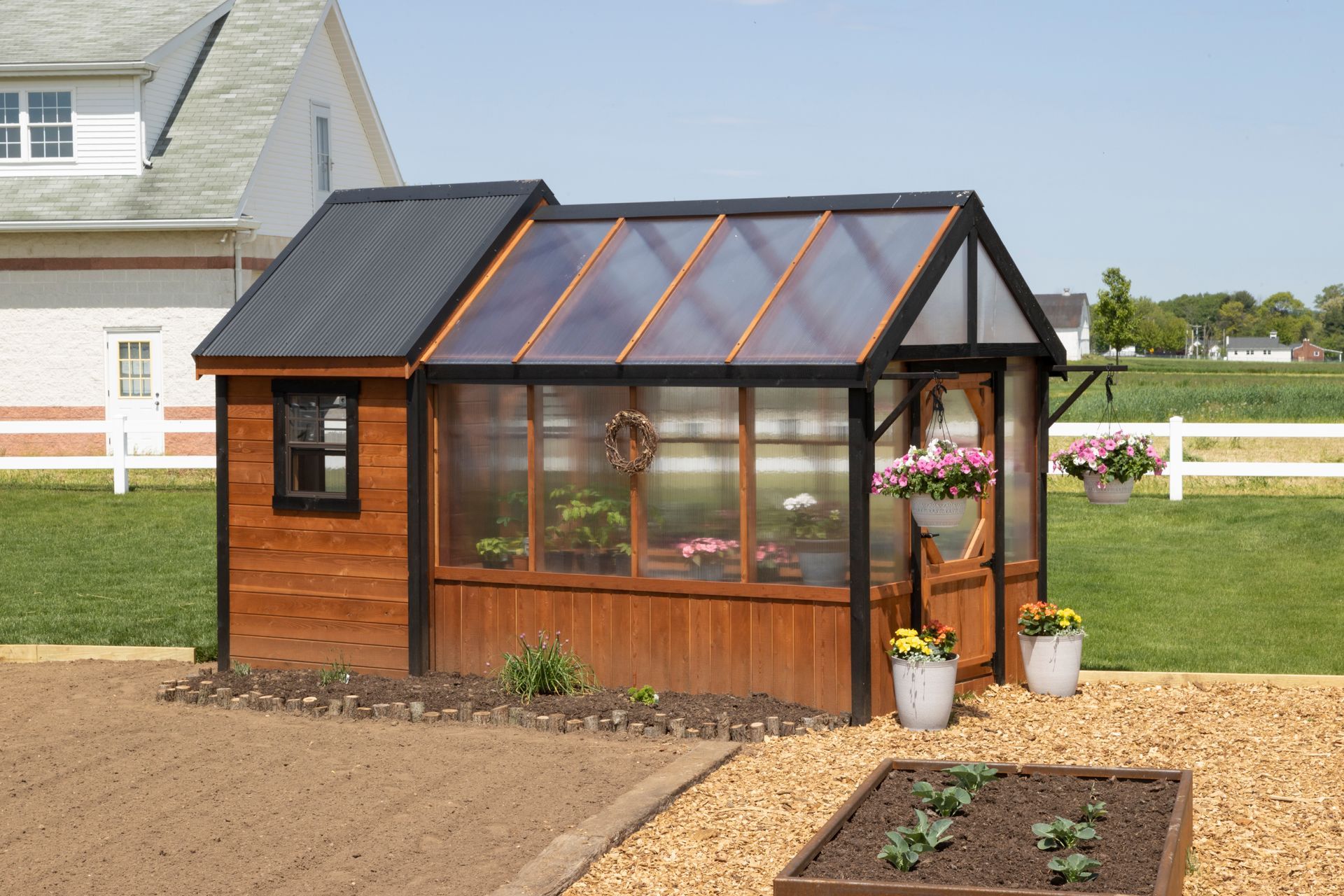 A wooden greenhouse with a dark metal roof and glass panels sits in a sunny backyard next to a raised garden bed.
