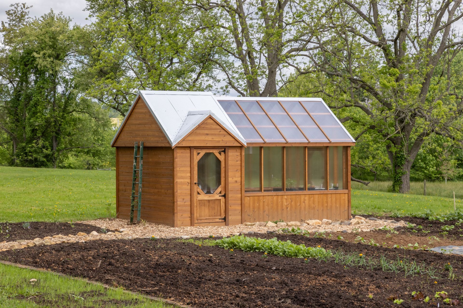 A wooden shed with a glass-paneled greenhouse wing, set in a garden with dark soil beds and mature trees in the background.