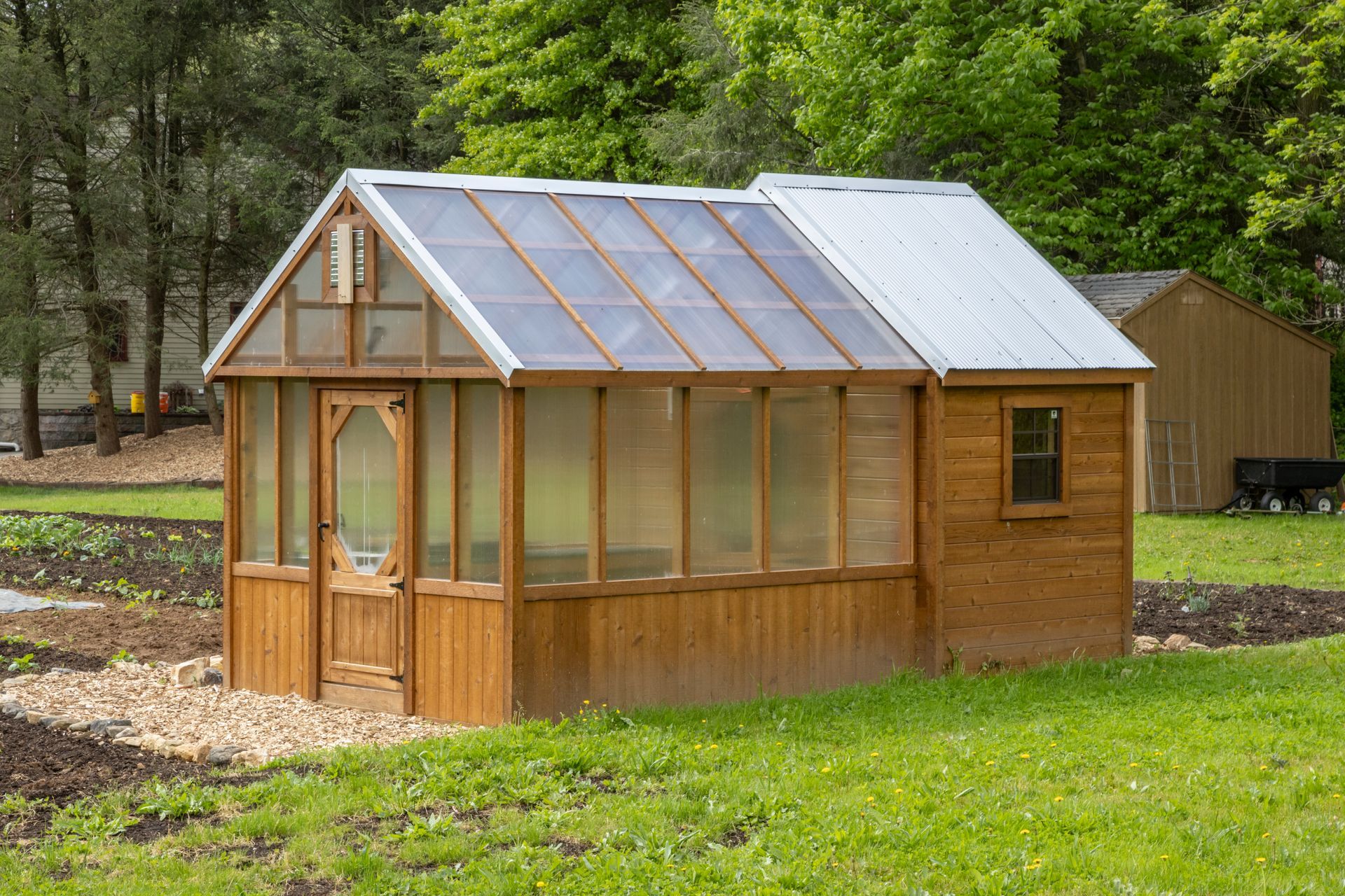 A wooden greenhouse with a glass roof and an attached shed sits in a grassy yard near a garden.