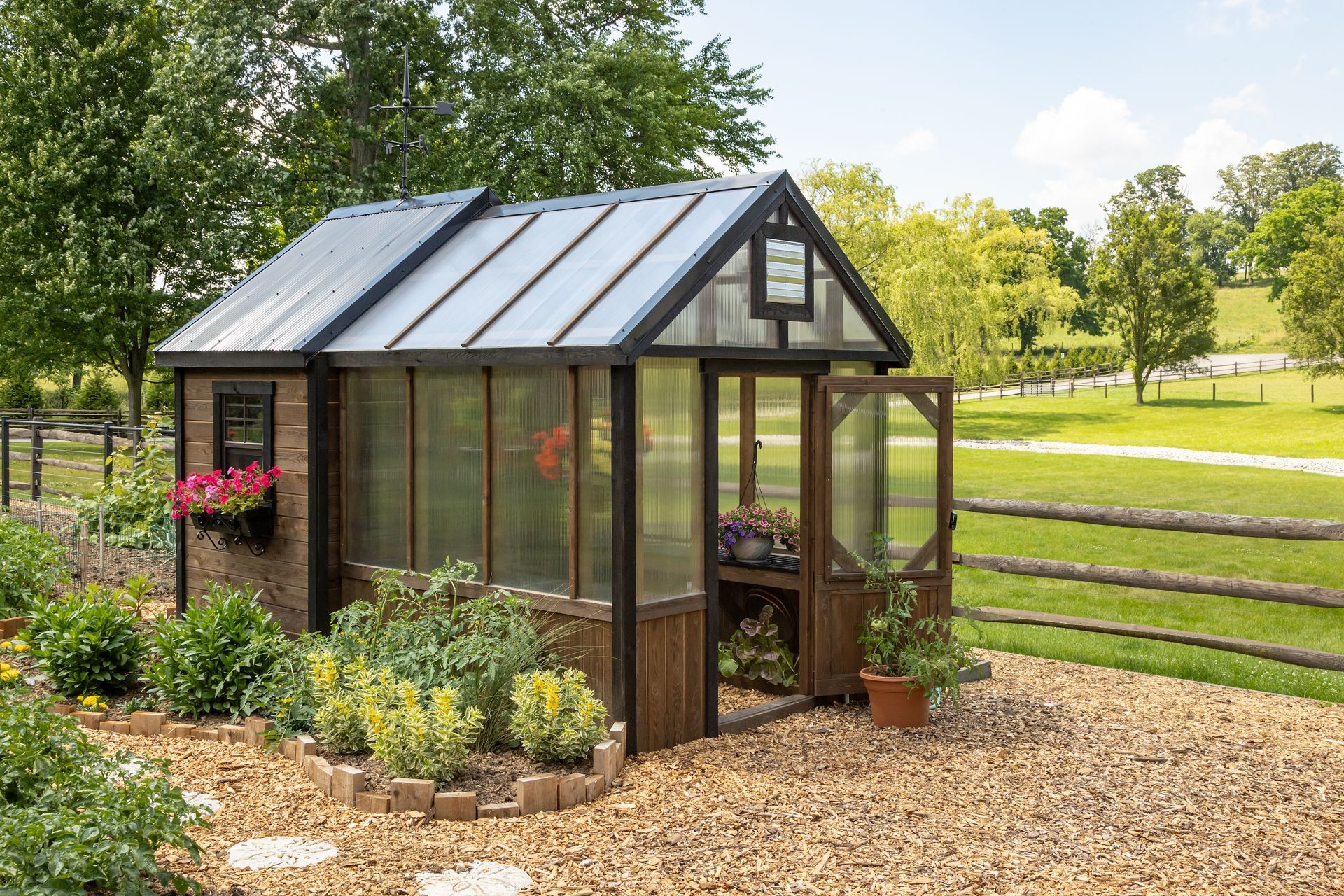 A small greenhouse with a dark frame and clear panels sits in a gravel garden, with green fields and trees beyond.