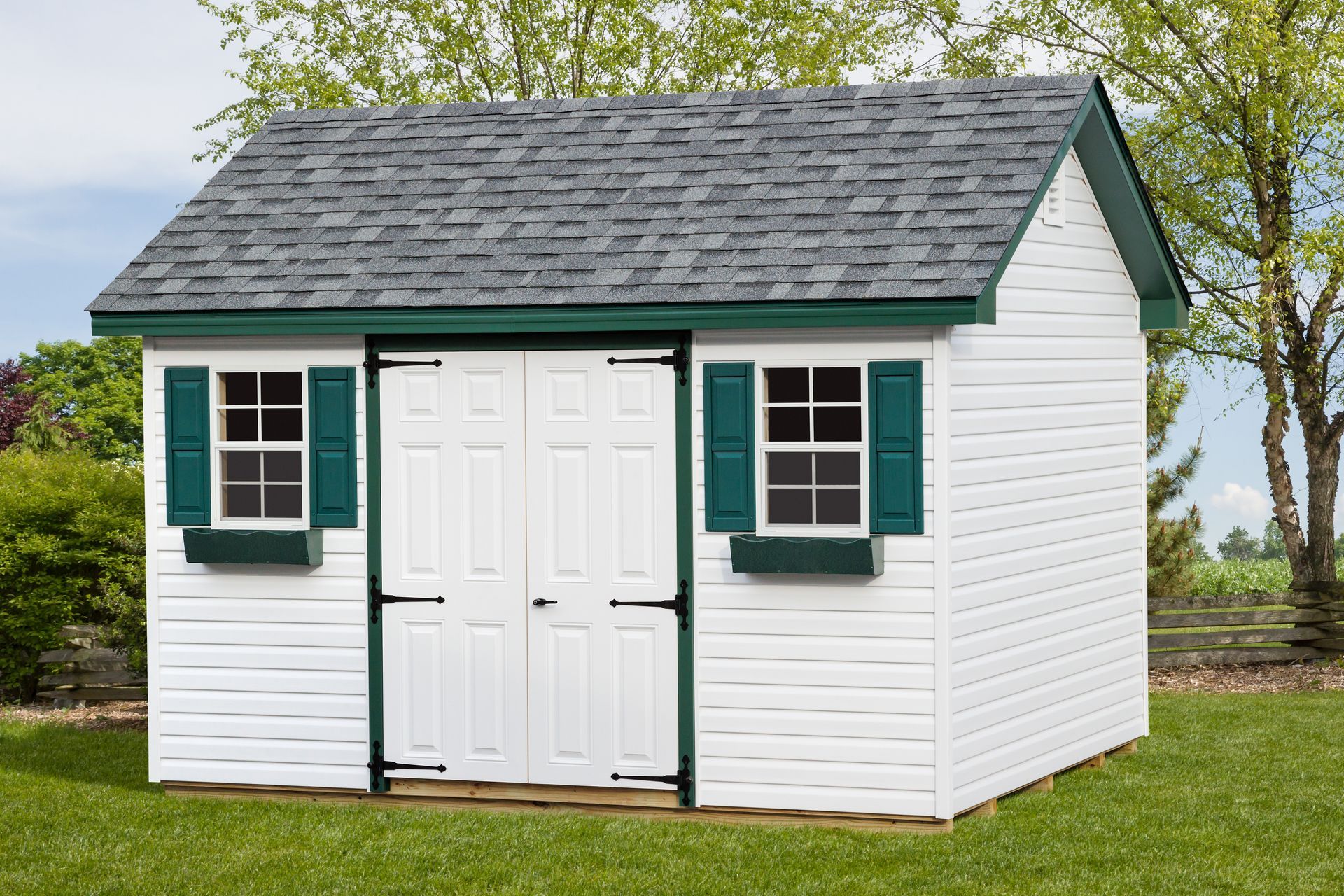 White shed with green trim, gray roof, and two windows with green shutters, set in a grassy yard.