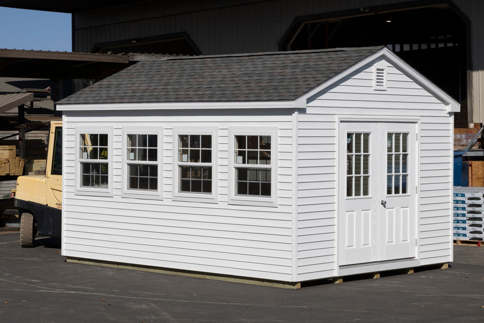 White shed with gray roof, windows, and a double door. Situated outdoors.