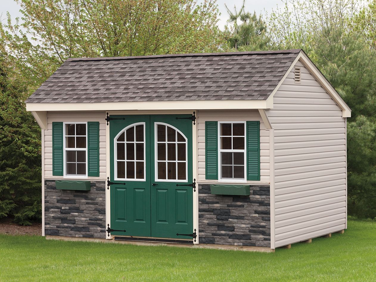 Tan shed with green doors, shutters, and stone facade, set on grass.