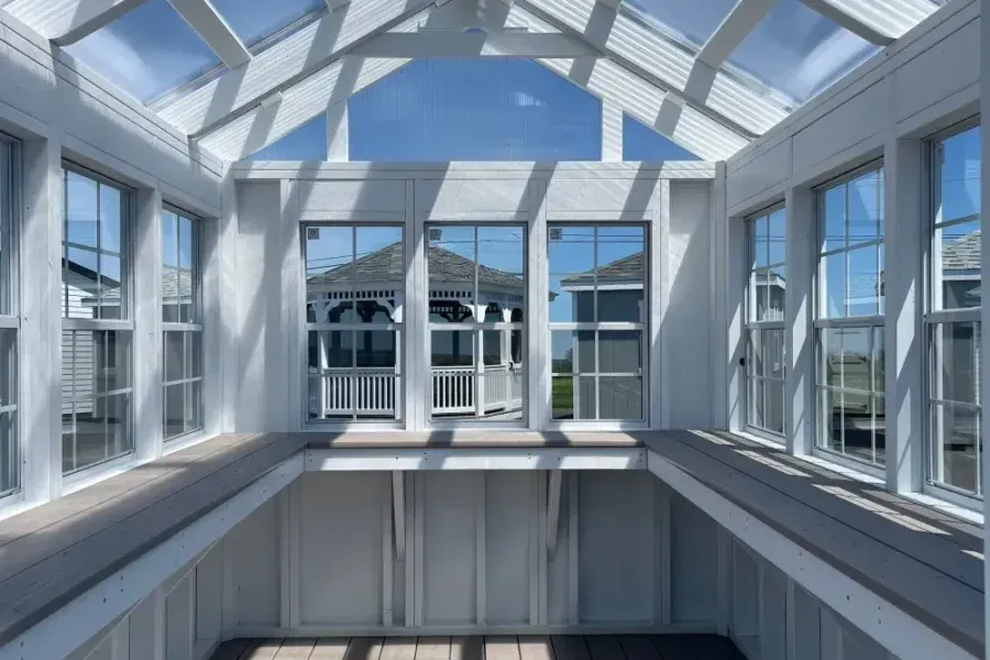A sunroom with white wooden framing, glass walls, and a glass-paneled roof, looking out toward a gazebo.