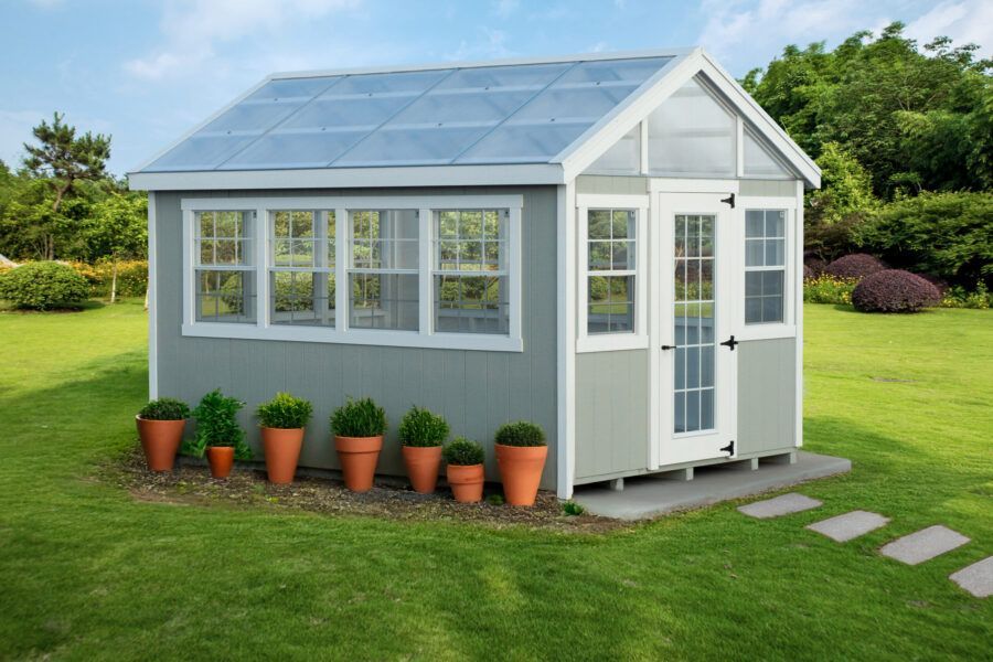 A gray greenhouse with a glass roof and white trim sits on a grassy lawn, with several potted plants lined up in front.