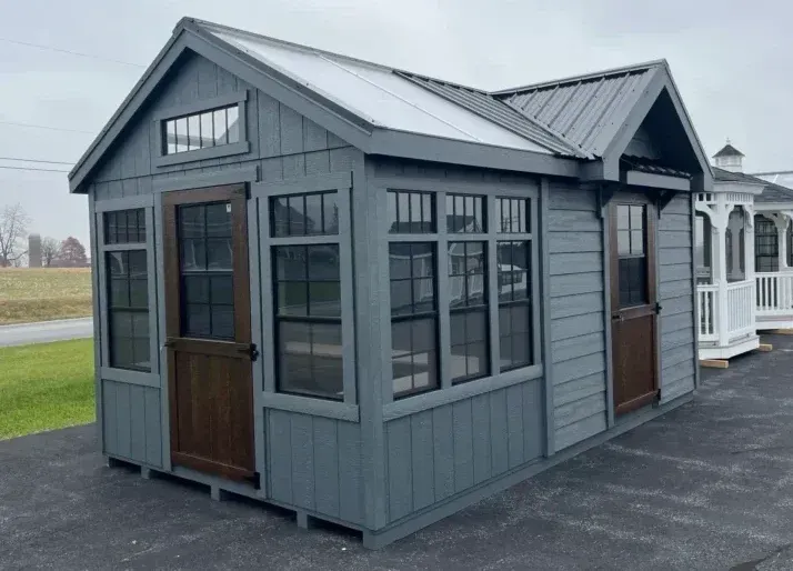 A grey garden shed with a metal roof, multiple windows, and two dark wooden doors, positioned on a paved lot.