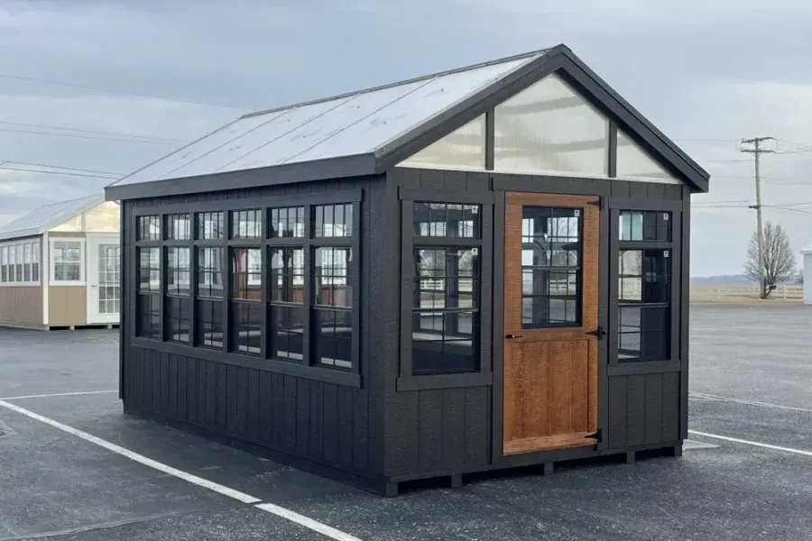 A dark gray greenhouse with a wooden door and a translucent peaked roof, sitting in an outdoor display lot.